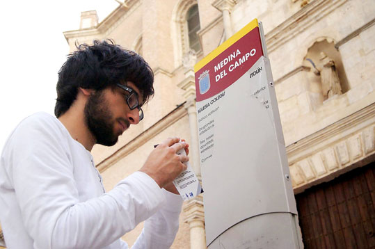 El estudiante medinense de ingeniería informática Adrián de la Rosa, promotor del proyecto, coloca un código QR en el cartel informativo de la Colegiata de San Antolín.